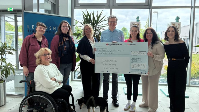 Eine Gruppe von acht Menschen, darunter eine Person im Rollstuhl und ein Hund, hält gemeinsam einen großen Spendenscheck in einem modernen Foyer hoch.