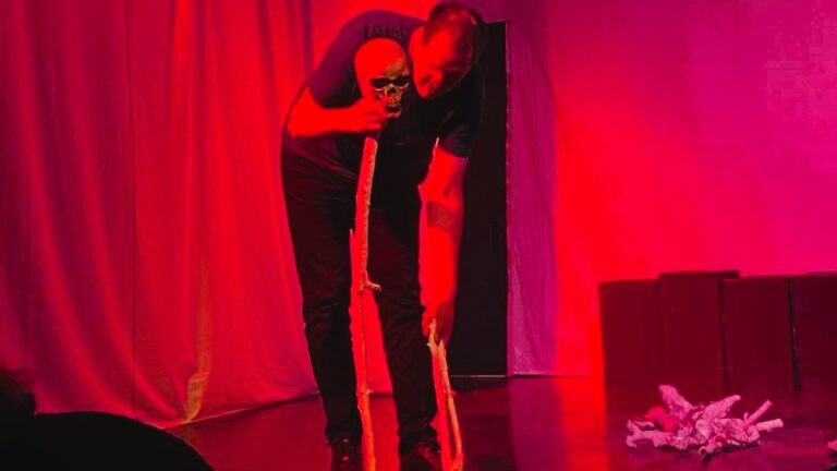 Eine Person steht gebückt auf einer Bühne im roten Licht und hält einen Totenschädel in der Hand